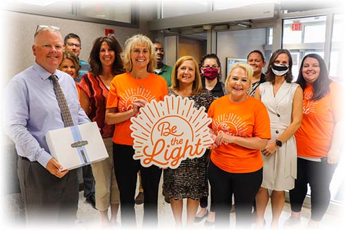 Group of people holding sign that says Be the Light in orange text on white background