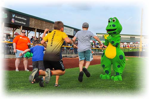 Two kids playing a game on a baseball field running towards frog mascot