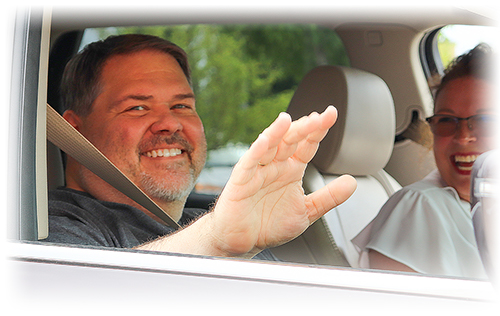 Dale smiling and waving out his car window. Wife Angie in the passenger seat smiling at Dale