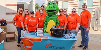Group of Ascentra staff standing behind blue Ascentra table at Clinton LumberKings game