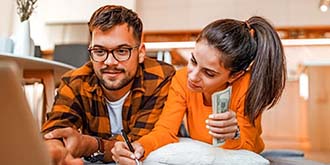 Couple laying on the floor budgeting on their laptop. Girl is a brunette with orange shirt and man has glasses, brown hair and wearing an orange and brown flannel