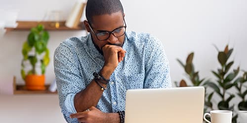 Man in blue shirt looking at his laptop with his hand to his mouth.