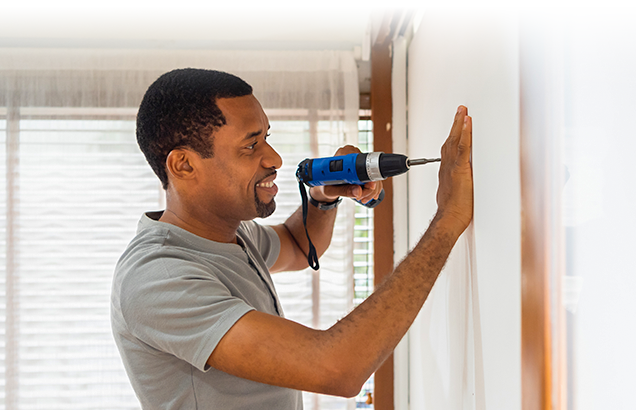 Man drilling hole into wall wearing a gray shirt and using a blue and black drill